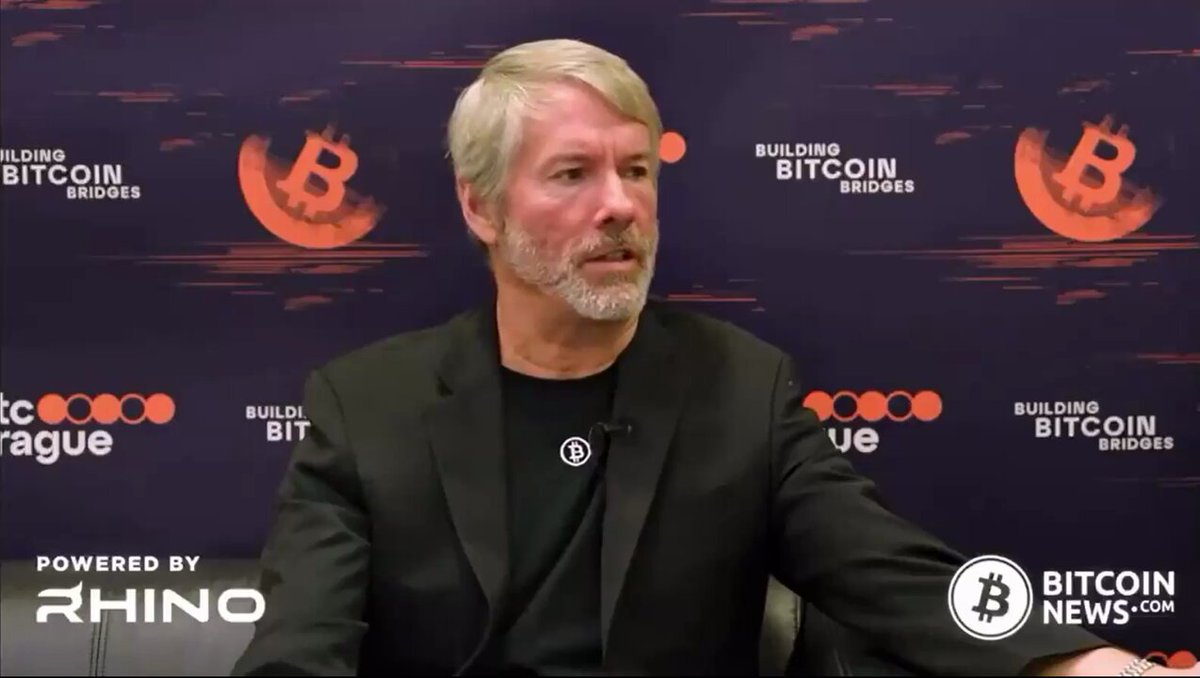 The image features a man sitting down, wearing a black suit jacket and tie. He appears to be talking about Bitcoin or blockchain technology during an interview. The background of the image includes a purple banner with logos for "Building Bitcoin Bridges" and "Rhinoo".