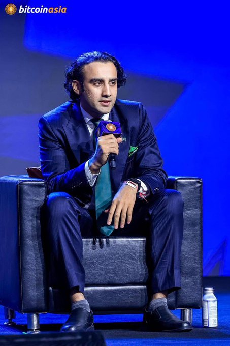 A man in a dark pinstripe suit with a teal tie speaking into a microphone while seated in a chair at the 'bitcoinasia' conference.