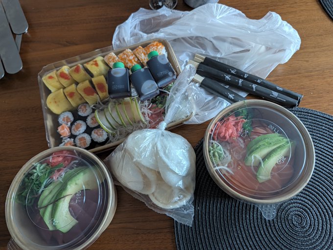 A table holds a sushi takeout spread with rolls, nigiri, sliced fruit, two bowls, soy sauce bottles, and three pairs of chopsticks. The extra chopsticks and family-style meal setup match the humorous caption about calling the kids to dinner in an empty house.
