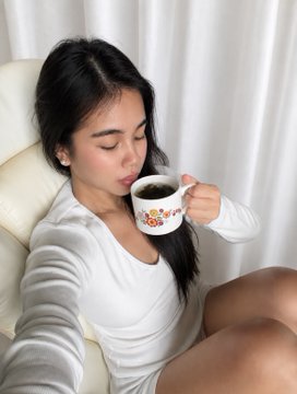 Selfie of a young woman with long dark hair wearing a white long-sleeve top, drinking from a decorative floral teacup while relaxing on a cream-colored chair.
