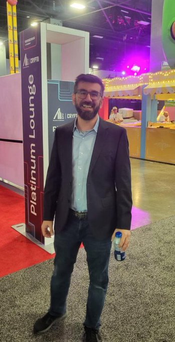 A smiling man wearing glasses, a dark blazer, and jeans stands in front of a "Platinum Lounge" sign featuring the "MJS Crypto" logo, holding a water bottle at what appears to be a crypto event. The image suggests his attendance at the event, reinforcing the caption's message about continued belief and participation in Pi Network despite skepticism.