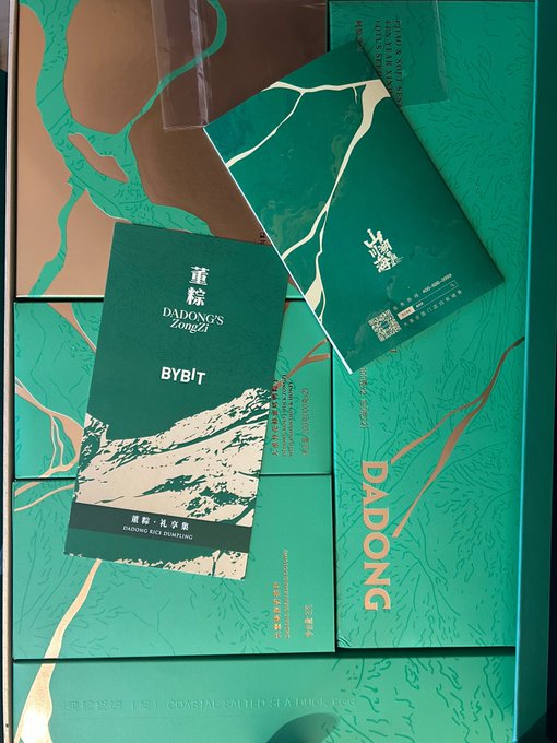 The image features a green box with gold lettering, which appears to be a gift or card from Bybit. The box contains two items - one being a small book and the other a larger envelope. These items are placed on top of a table, surrounded by various other cards and envelopes. The post in question is about a product called "Bybit", which seems to be related to these cards and envelopes.