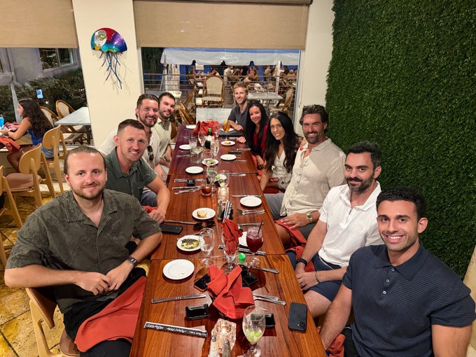 A group of ten smiling colleagues pose together around a long dining table at a restaurant, with place settings, drinks, and a red napkin visible in front of them. The scene reflects the celebratory team dinner mentioned in the post, marking nearly one year since the launch of ThirdLayer and acknowledging the core team behind it.