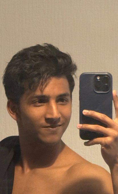 A young man with dark wavy hair takes a mirror selfie with a navy iPhone, shirtless with a towel draped over one shoulder, smiling subtly as he greets the day. The casual "GM" (Good Morning) post reflects the relaxed, easygoing mood of the selfie, suggesting a refreshing start to the Wednesday countdown toward the weekend.