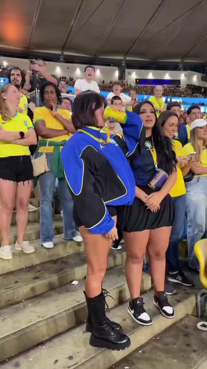 The image shows two women standing together, one wearing black boots and a blue jacket with white trim, while the other wears a yellow shirt. They are surrounded by a crowd of people who appear to be watching them intently. The background reveals that they are in an indoor sports arena or stadium, possibly attending a sporting event.