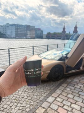 The image features a person holding a cup of coffee, with a car parked nearby. The scene takes place near a body of water, possibly a river or a lake, which adds an interesting element to the composition. In the background, there are several buildings visible, suggesting that this location might be in a city or urban area. The image also contains text elements such as "zolana summer kick off in Berlin was great to share the stage with @yesjess and chat about privacy on Solana & why it matters also huge shoutout to @pattiruss_ @zCase_ & the whole @SuperteamDE team for organizing this mini breakpoint in our home country only higher".