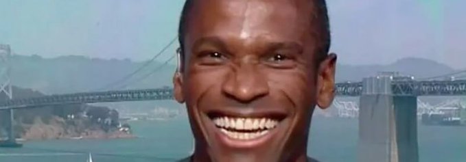 A smiling man with a wide grin is shown against a backdrop of the San Francisco Bay Bridge and water.