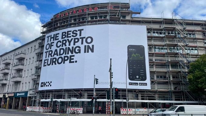A massive billboard on a building wrapped in scaffolding advertises "THE BEST OF CRYPTO TRADING IN EUROPE" for OKX, with downloads available on the App Store and Google Play, seen alongside the accompanying Instagram caption celebrating OKX's expanding presence across European neighborhoods.