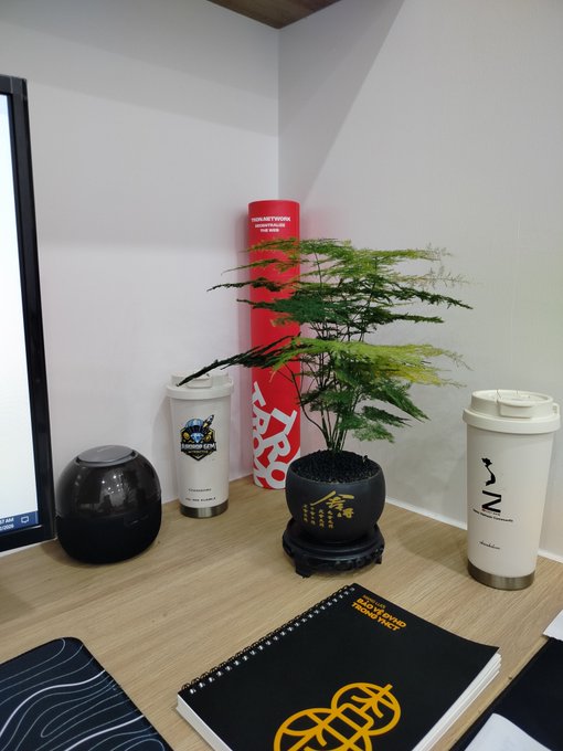 A desk setup featuring a bonsai plant in a black pot with a Chinese character, a red cylindrical item with Asian text, a 'FIGHTER GYM' tumbler, a white 'INZ' tumbler, and a notebook with a gold knot design.