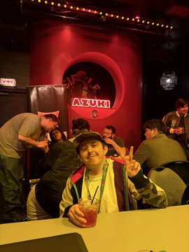 A man wearing a baseball cap and lanyard flashes a peace sign while holding a drink at a table, with a sign reading "AZUMI" visible in the background at what appears to be the Anime Expo. The image captures the casual, social atmosphere of the convention's day 0 events, likely taken in a restaurant or bar area associated with the expo.