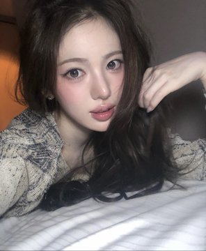 A close-up selfie of a young woman with long dark hair, wearing a denim jacket and heavy blush, lying on white bedding.