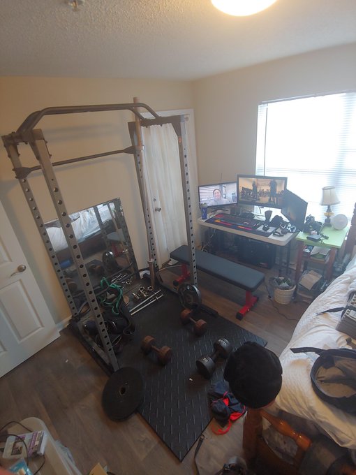 A home gym setup features a power rack with a mirror, a bench, dumbbells, a weight plate, and a desk with monitors in the background.