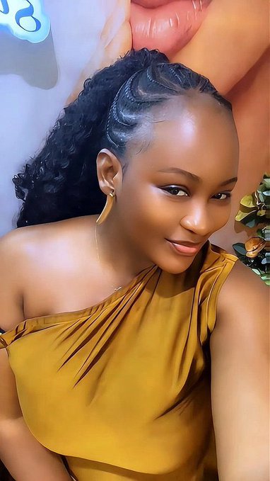 A smiling woman with a curly ponytail poses in a mustard yellow off-shoulder top, wearing a unique leaf-shaped earring, with what appears to be a ring light reflection in the upper corner.