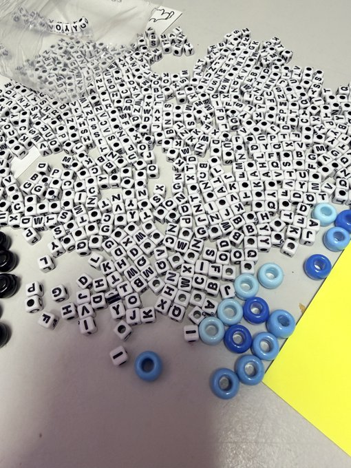 A massive pile of white plastic alphabet beads dominates the scene, accompanied by scattered blue round beads and a yellow sheet in the background. The post's note about "running out of e's" connects to the image, which shows beads nearly depleted of the letter "E," highlighting the challenge of assembling words or a full alphabet set.