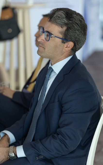 A man with grey hair and glasses, wearing a dark navy suit with a light blue shirt and patterned tie, seated in profile. Another person is partially visible behind him.