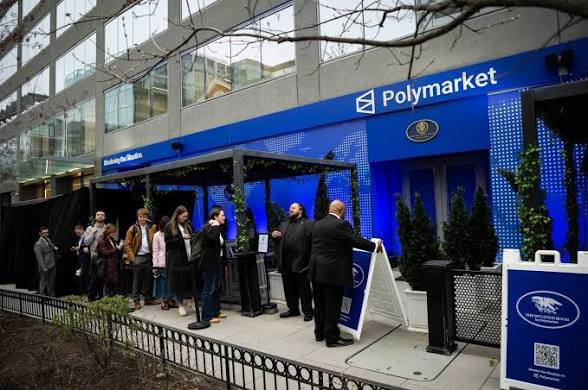 An outdoor storefront for Polymarket featuring blue branding, with a group of people gathered near the entrance and a sign mentioning "The Pennsylvania Gaming Control Board."