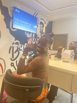 A woman wearing sunglasses on her head and a brown tank top sits in an office chair, smiling and posing for the camera, with a wall behind her featuring a monitor that reads 'THE ECONOMICS OF FOR THE INTERNET.'