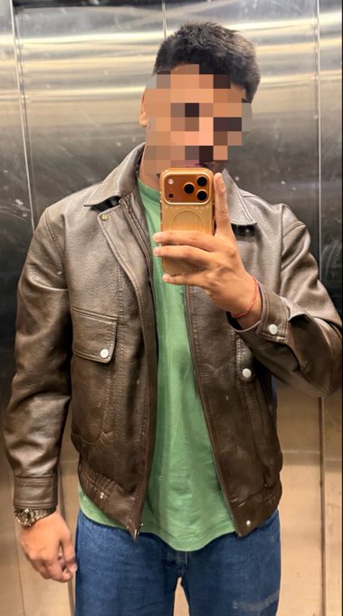 A man taking a mirror selfie in an elevator wearing a brown leather jacket over a green t-shirt and blue jeans, with his face blurred and holding a gold smartphone.