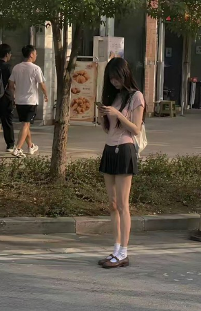 A young woman stands on a sidewalk looking down at her phone, wearing a pink top, dark pleated skirt, white socks, and brown Mary Jane shoes, with two men walking away in the background near a menu board.