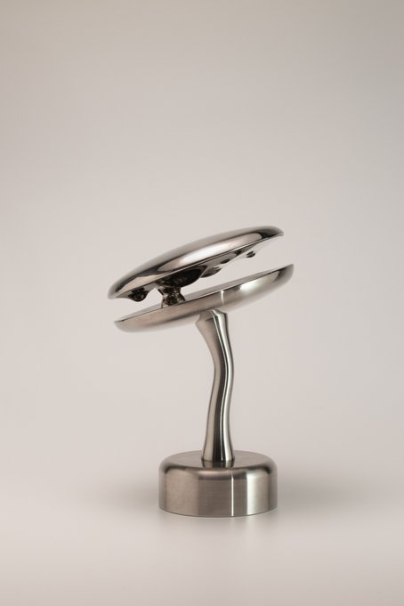 A chrome-plated metallic object with a curved, twisted stem and circular top mounted on a cylindrical base, photographed against a plain off-white background.