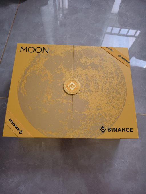 A golden Binance-branded "MOON" gift box sits on a gray tiled floor, featuring the iconic Binance logo on a central wax-seal-style emblem and a printed moon surface design. The packaging ties to the post's celebration of Binance's Mid-Autumn Festival merchandise, which the user praises for its aesthetic quality and rumored $100 price tag.