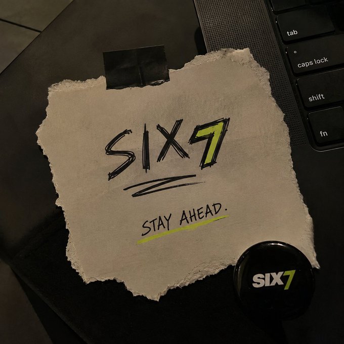 A torn piece of paper taped to a desk next to a laptop keyboard reads "SIX7 STAY AHEAD." in handwritten-style text, accompanied by a matching black button.