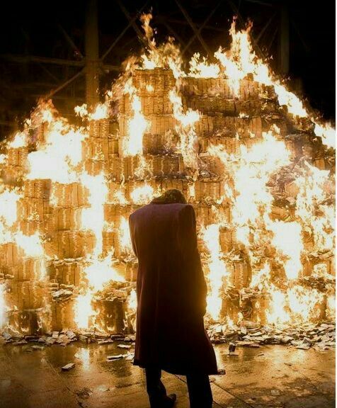 A dramatic nighttime photograph showing a figure in a long coat standing with their back turned, facing a massive, towering structure of stacked cardboard boxes engulfed in bright orange flames.