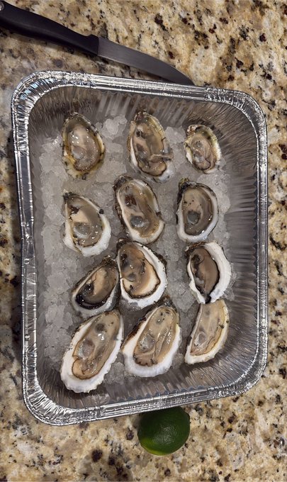 A tray of fresh oysters on ice with a lime wedge, placed on a granite countertop next to a knife.