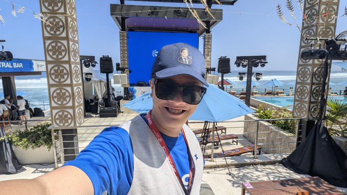 A selfie of a man wearing a blue baseball cap, sunglasses, and a lanyard, taken at an outdoor beachside venue with a stage, pool, and ocean visible in the background.