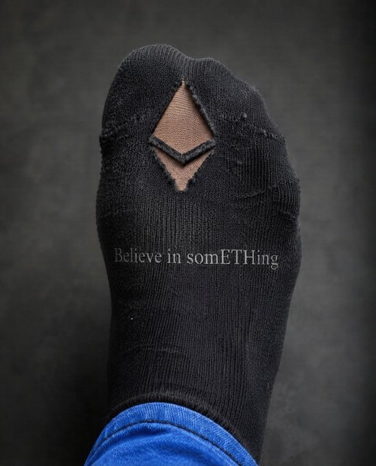 A worn black sock with a hole exposing a toe, displaying the text "Believe in somETHing" in a Nike-style font, cleverly riffing on the iconic slogan to celebrate Ethereum's birthday in meme format.