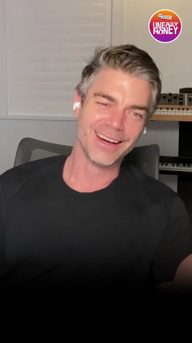 The image features a man wearing a black shirt sitting in front of a computer screen with his head tilted back while smiling at the camera. The background includes a window, suggesting that he might be working from home or in an office setting. There is also a keyboard and mouse visible on the desk next to him.