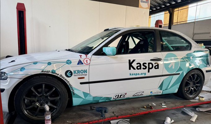 A white race car with teal geometric graphics parked in a garage, displaying 'Kaspa' branding with the URL 'kaspa.org' along with 'KRON technology' and 'GT2' logos.