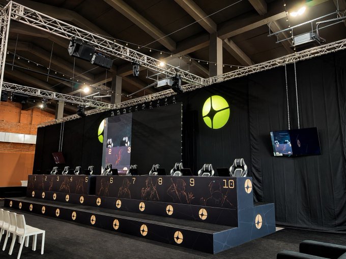 An empty gaming setup features a tiered desk with ten gaming chairs, screens, and a large yellow logo on a black backdrop, suggesting an esports event venue.