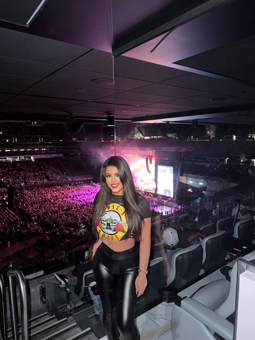 A smiling young woman with long dark hair stands in the upper seating area of a large arena during what appears to be a Guns N' Roses concert, wearing a cropped graphic t-shirt and black pants.