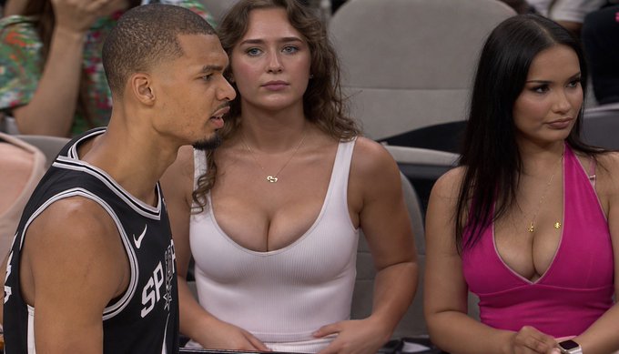 The image shows a group of three people sitting together, with one person wearing a black and white jersey while another wears a pink dress. The third person appears to be looking at something or someone off-camera. There are several other people visible in the background, but they do not seem to be the main focus of the photo.