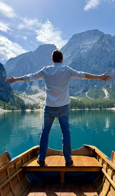 A person in a striped shirt and jeans stands on a wooden boat with arms outstretched, facing a vibrant turquoise lake surrounded by tall mountains.