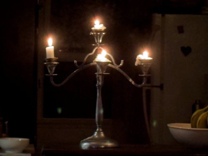 A dimly lit photograph showing a silver candelabra holding four lit candles, with a bowl of bananas visible to the right.