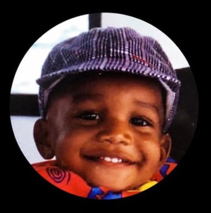 A circular-cropped vintage photograph shows a smiling young child wearing a striped flat cap and a colorful patterned shirt.