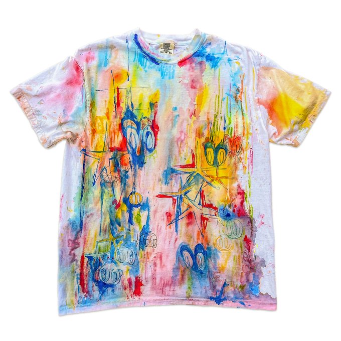 A colorful tie-dye style t-shirt featuring a hand-painted artistic design with cartoon-like characters and vibrant splashes of pink, blue, yellow, and red paint.