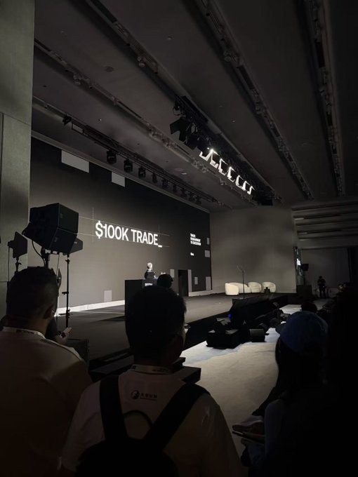 An audience watches a presentation at a '$100K TRADE' event in a darkened auditorium with large projection screens on stage.