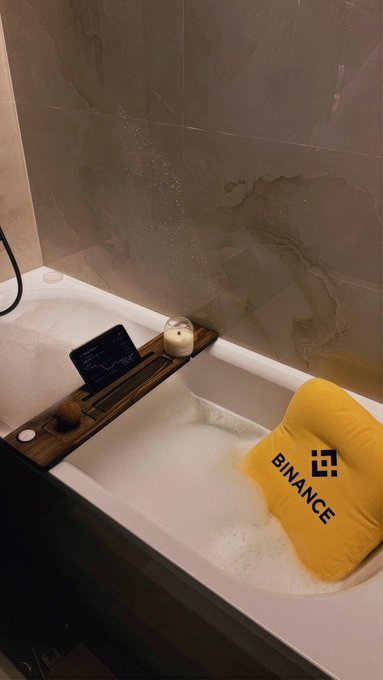 A bubble bath scene features a yellow pillow branded with the "BINANCE" logo and the BNB-style icon floating in the tub, alongside a phone displaying a chart and a lit candle, suggesting a stressed crypto trader trying to unwind after a tough market week. The Binance pillow with its logo directly nods to the crypto exchange amid the post's weary "What a week, huh?" sentiment.