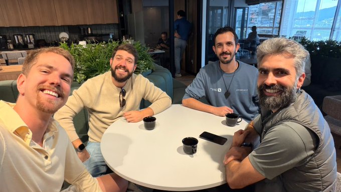 Four men smile at the camera while seated around a small round white table in a modern café or co-working space, with two black coffee cups and a smartphone placed in the center. The relaxed group photo aligns with the post's caption about sharing lunch with builders during a visit to Floripa (Florianópolis), Brazil, with the man on the right wearing a hoodie that appears to display partial text/logos consistent with the @zanon1guilherme and other tagged builders.