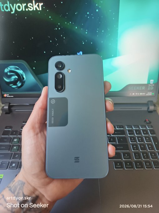 A hand displays the back of a blue/gray Solana Seeker smartphone featuring the "SEED VAULT" branded panel and "M" logo, held up in front of a laptop displaying the Seeker homepage. The post invites other Solana Mobile Seeker owners to follow back, showcasing this device as an example of the community's mobile hardware.