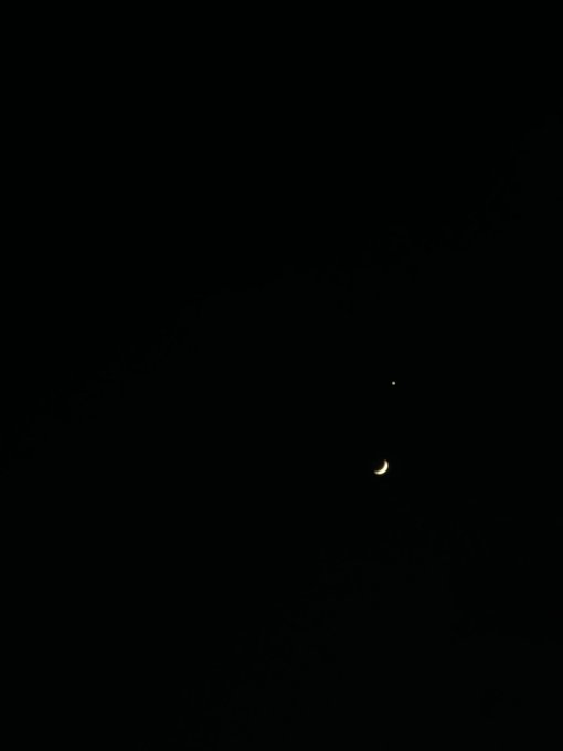 A dark night sky featuring a thin crescent moon in the lower right and a small point of light (likely a planet) above it.