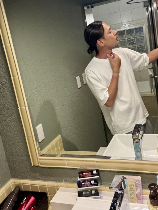 A person in a white t-shirt stands in a bathroom mirror, looking up while touching their neck, with various toiletries visible on the counter below.
