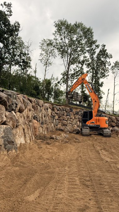 An orange Doosan excavator is actively placing large boulders along a hillside to construct a retaining wall on a dirt worksite. The image illustrates the early construction phase referenced in the caption, where "Stoney" has nearly finished building this wall before moving on to stack another 60k-handle boulder structure.