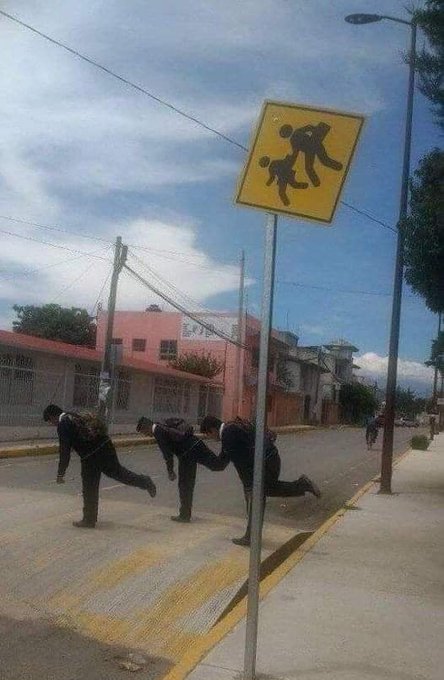 A group of people walking down a street in a single-file line, mimicking the pose of the yellow pedestrian crossing sign above them.