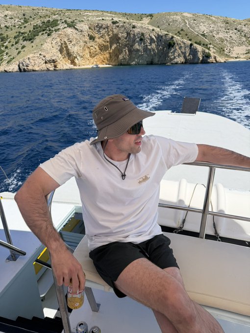 The image features a man sitting on a boat, wearing sunglasses and a hat. He has a beer in his hand, which he appears to be enjoying while looking out at the water. The background of the photo shows a beautiful view of the ocean with mountains in the distance. There is also an advertisement for a website called HyperliquidX visible in the image.