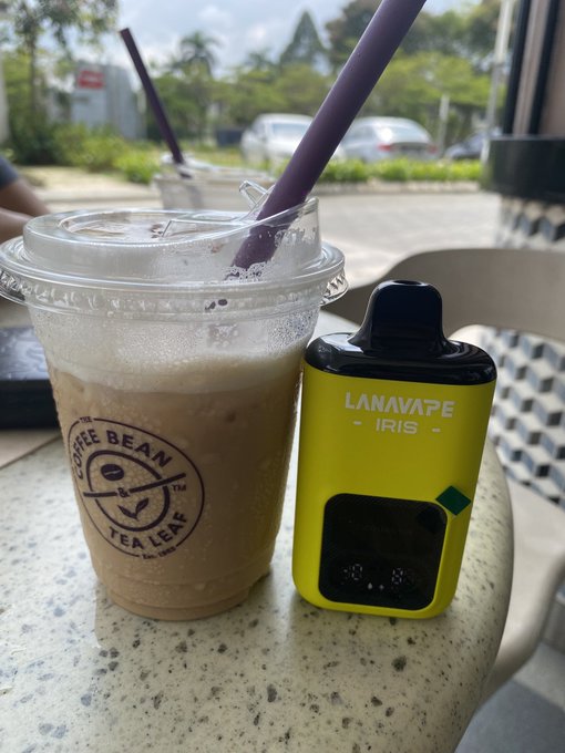 An iced beverage from 'The Coffee Bean & Tea Leaf' in a clear plastic cup with a purple straw, placed next to a yellow 'LANAVAPE IRIS' vape device on a speckled table.