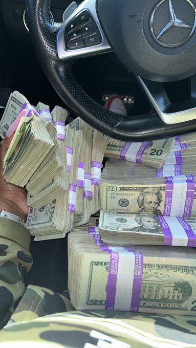 A person's hand holds stacks of $20 bills wrapped in purple currency bands beside a Mercedes-Benz steering wheel, while the accompanying text's reference to "greens" and "Claim it" suggests an aspirational message about accumulating wealth.